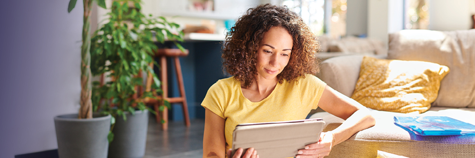 Woman sitting on couch reading a tablet in a bright living room with plants nearby